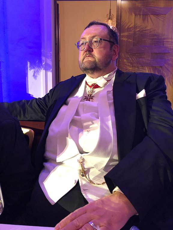 A man with vest, jacket, pocket square and glasses sits leaning next to a window, in the foreground his ringed hand