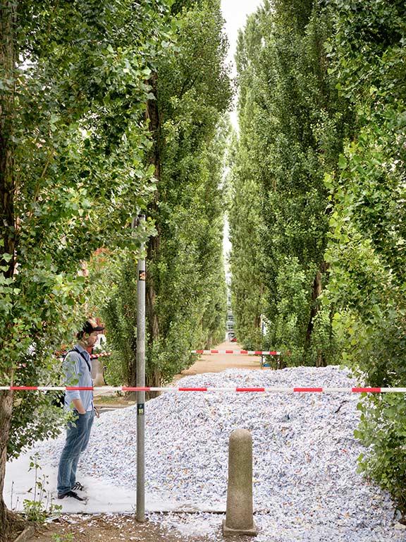 A man with a baseball cap and his hands in his pockets, in profile on the left, stands in front of a large pile of paper scraps, in the foreground a red and white barrier tape, to the side and in the background trees and a reddish gravel road