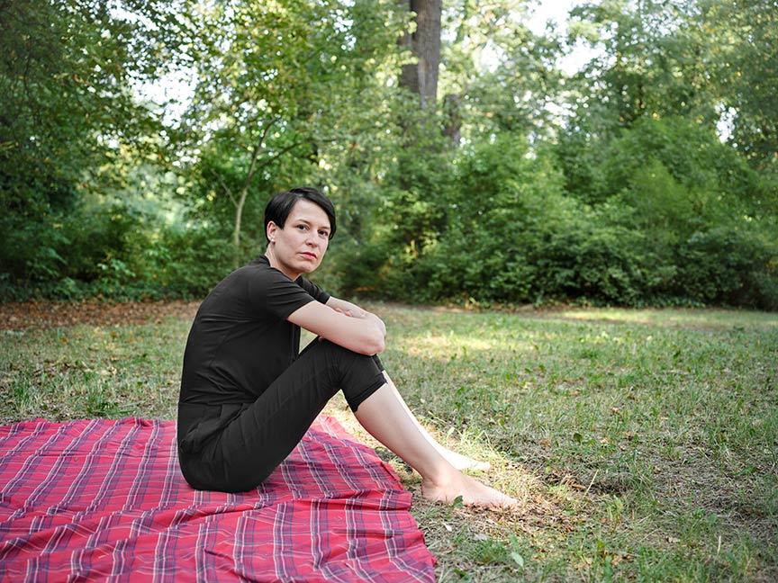 Eine schwarz gekleidete Frau mit kurzen Haaren sitzt auf einer roten Picknickdecke auf einer Wiese und schaut in die Kamera