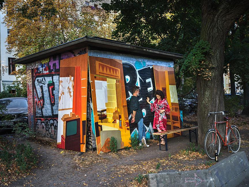 Ein Mann in Jeans, Caro-Hemd und Turnschuhen und eine barfüßige Frau im bunten Kleid stehen einander vor einem bunt bemalten Schuppen gegenüber, vorne rechts lehnt ein Fahrrad an einem Baum, auf dem Boden davor steht eine Bierflasche