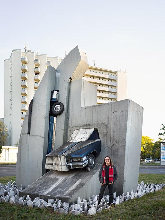 Ein grau-schwarz gekleideter Mann mit rotem Schal steht vor einer Skulptur, bei der ein halbes Auto schräg aus einer Betonwand hervorkommt, im Hintergrund ein hoher Wohnblock