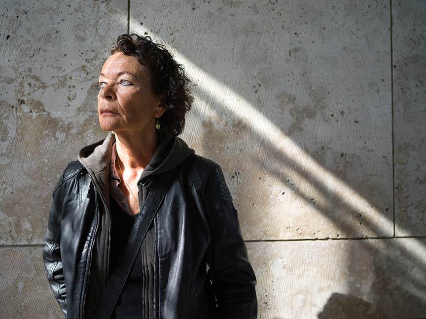 A woman in black leather jacket stands in front of a gray-brown wall and looks to the side