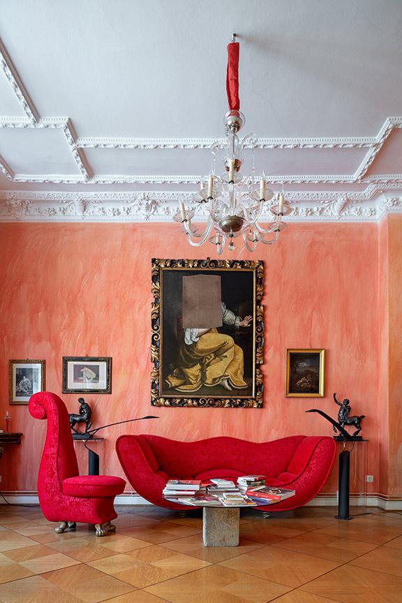 View of a room with stucco ceiling, chandelier, red designer couch set and above the sofa on the reddish painted wall a gold framed old oil painting of a woman with a rectangular piece of canvas missing at the level of the head