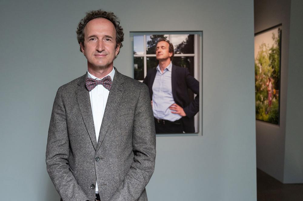 A man in a suit and bow tie stands smiling in an exhibition in front of a portrait of himself on the wall with his eyes closed and his hands resting at his sides