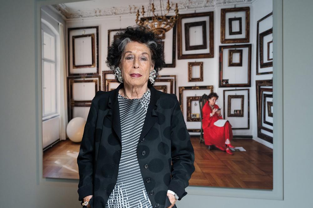 An elderly woman with large earrings and her hands in her pockets stands in front of a portrait painting of herself sitting in a chair in a room with many empty frames on the walls