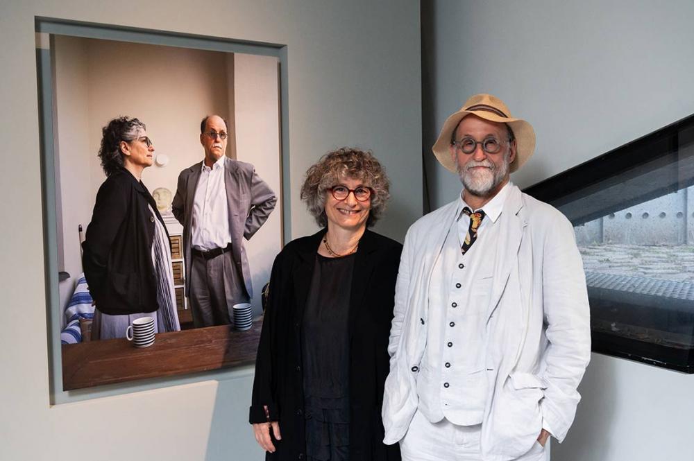 Ein lächelndes Paar, beide grauhaarig, sie ganz in schwarz, er im weißen Anzug und mit Strohhut steht neben einem Porträt von ihnen in der Ausstellung, auf dem Porträt stehen sie hinter einem Tisch und schauen in unterschiedliche Richtungen