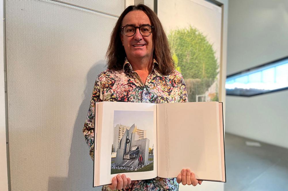 Ein Mann mit langen Haaren, Brille und buntgemustertem Hemd steht in einer Ausstellung und hält ein aufgeschlagenes Buch in die Kamera, in dem ein Porträtfoto von ihm ist