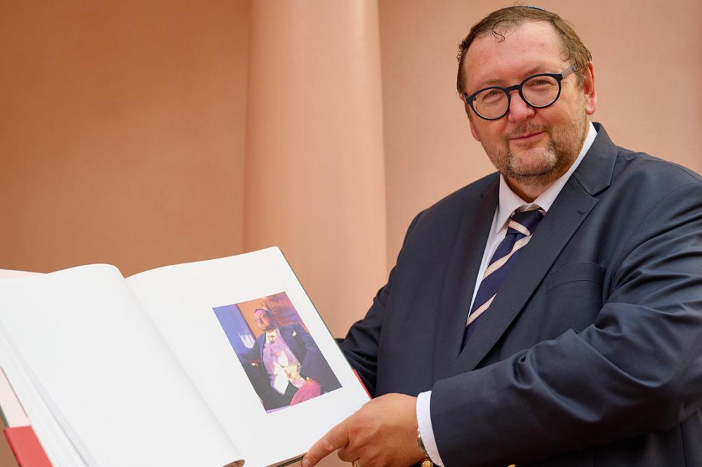 A man in a suit holds an open book into the camera, in the book is a portrait of him