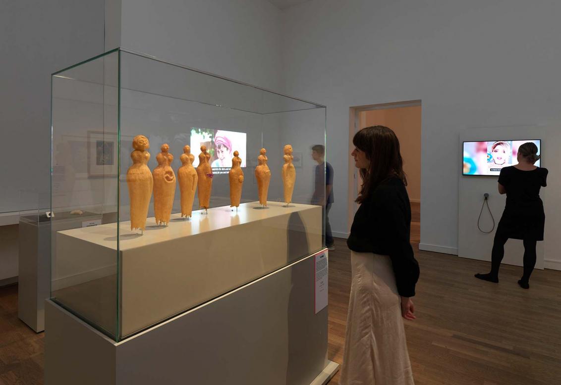Exhibition view, a woman stands in front of a display case containing orange figures.
