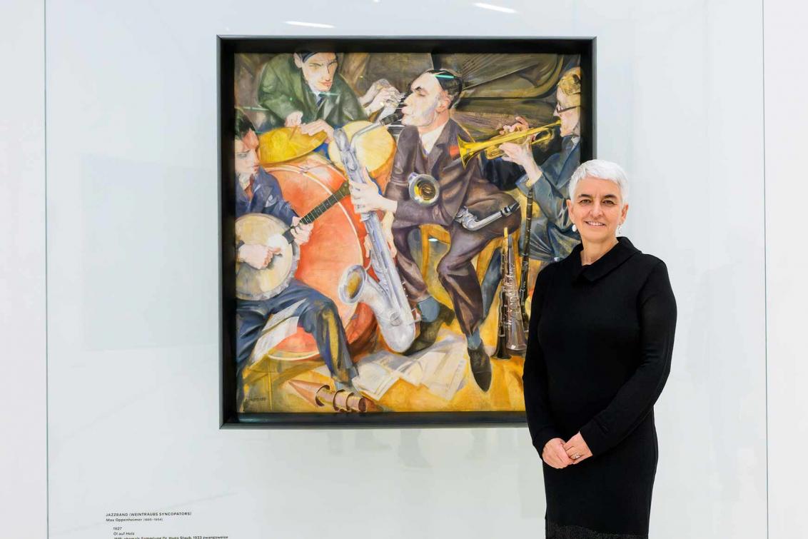 A woman dressed in black with short white hair in front of a cubist-inspired painting depicting a jazz band.