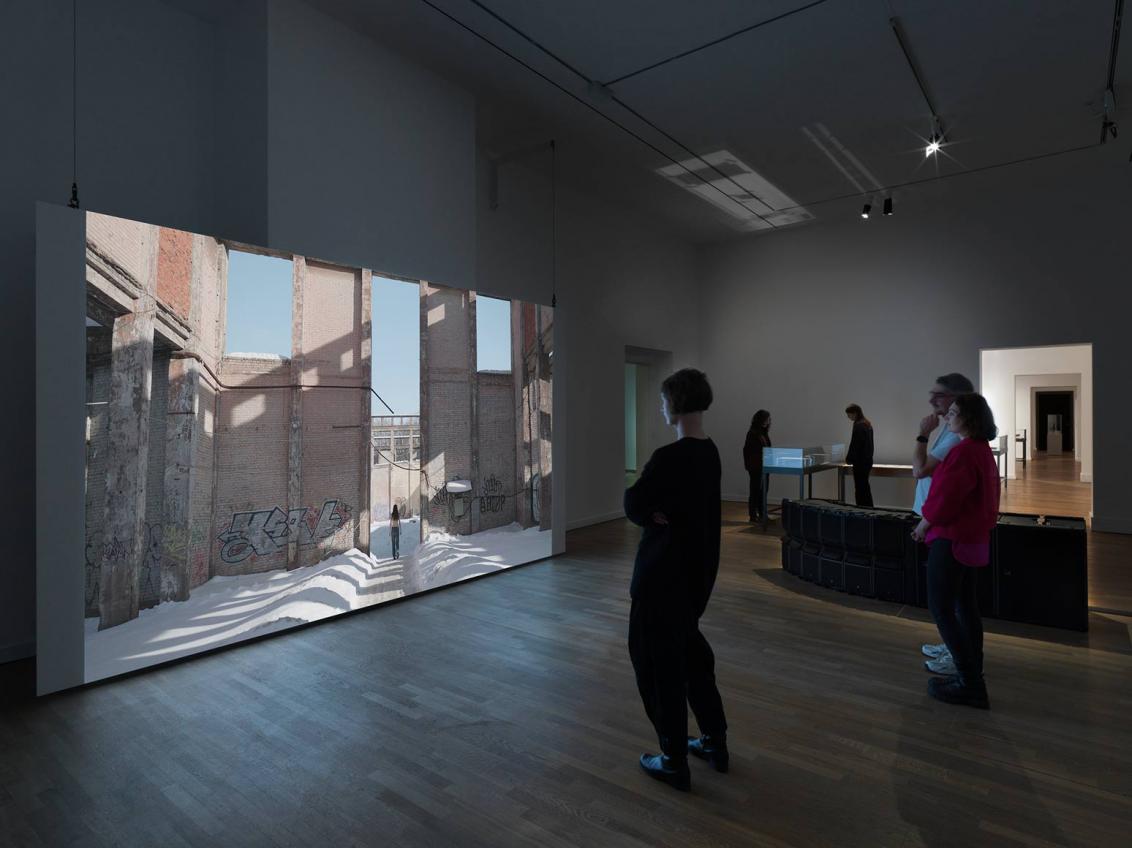 Visitors look at a large screen on which a long-haired person wearing jeans and a bare upper body is walking through the snow in a dilapidated brick building.