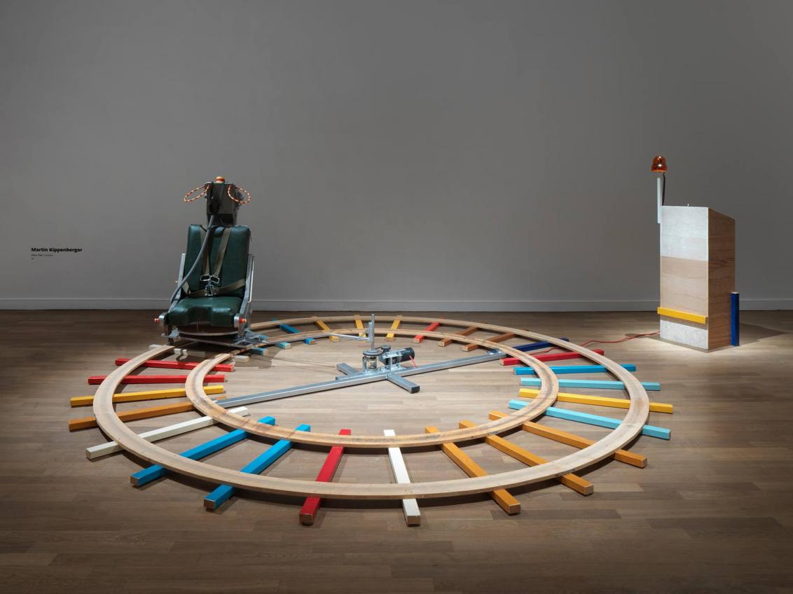 Exhibition room with colorful wooden rails running in a circle on the floor, with an ejection seat on top and on the right a wooden desk with an orange light wired to the seat.