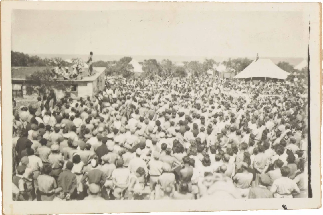 In the black-and-white photo taken from diagonally above, a large crowd of people is facing a house on whose roof a man is standing and speaking to the crowd.