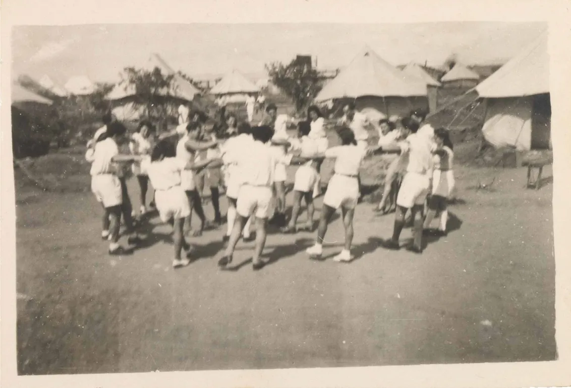 In the black and white photograph, men and women in white shorts and white blouses are dancing in an inner and an outer circle, holding hands and swinging out with their left foot. White tents can be seen in the background.