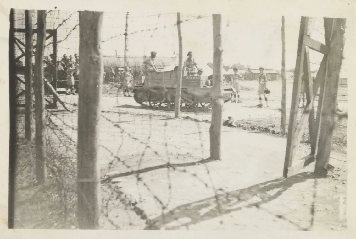 A barbed wire fence attached to wooden pillars can be seen in the foreground of the black and white photo. In the background are two tanks with soldiers sitting on them and a few internees standing around them.