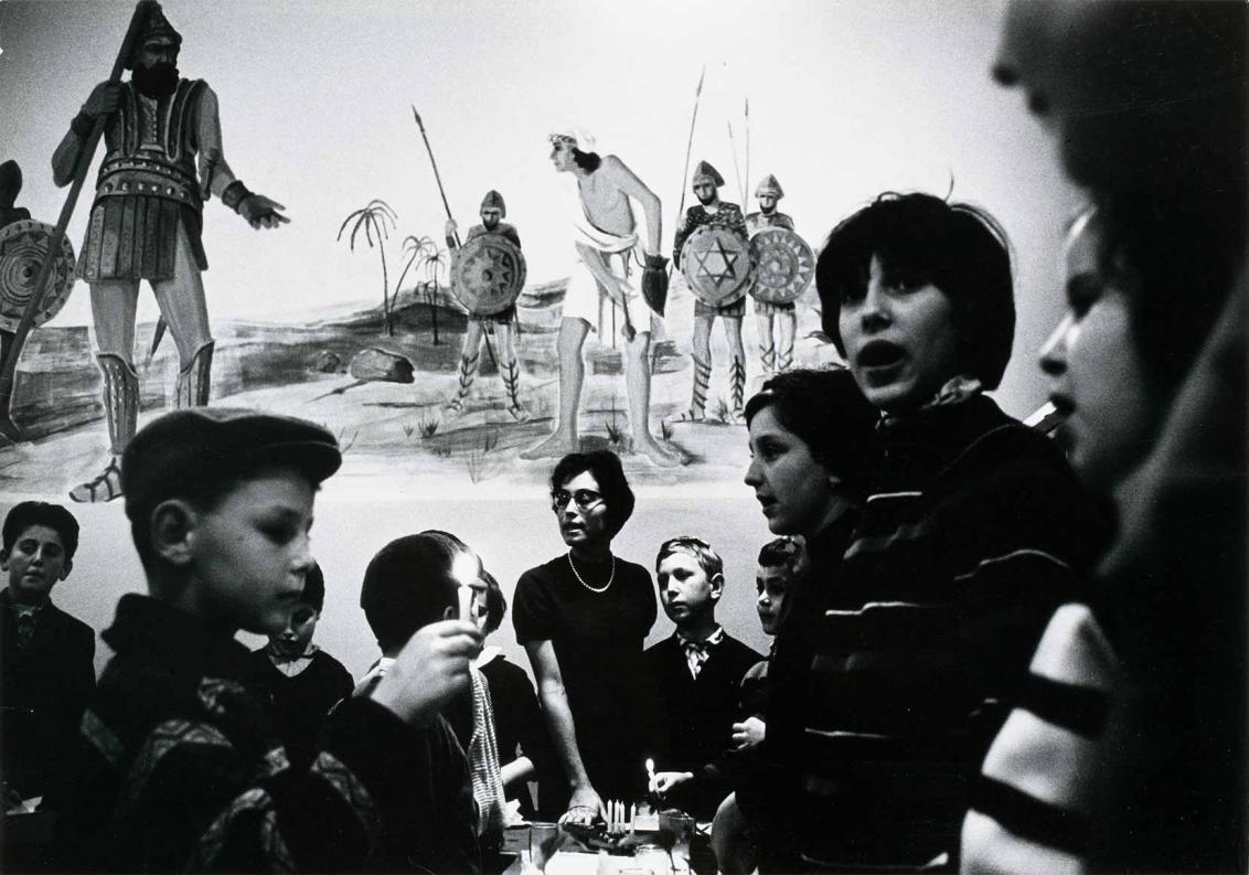 Schwarzweiß-Fotografie: Das Foto zeigt eine Dame und mehrere Kinder, zwei der Kinder halten eine Kerze in der Hand. Sie befinden sich in einem Raum und umringen einen Tisch. Auf der Wand im Bildhintergrund ist der Kampf von David gegen Goliath dargestellt.