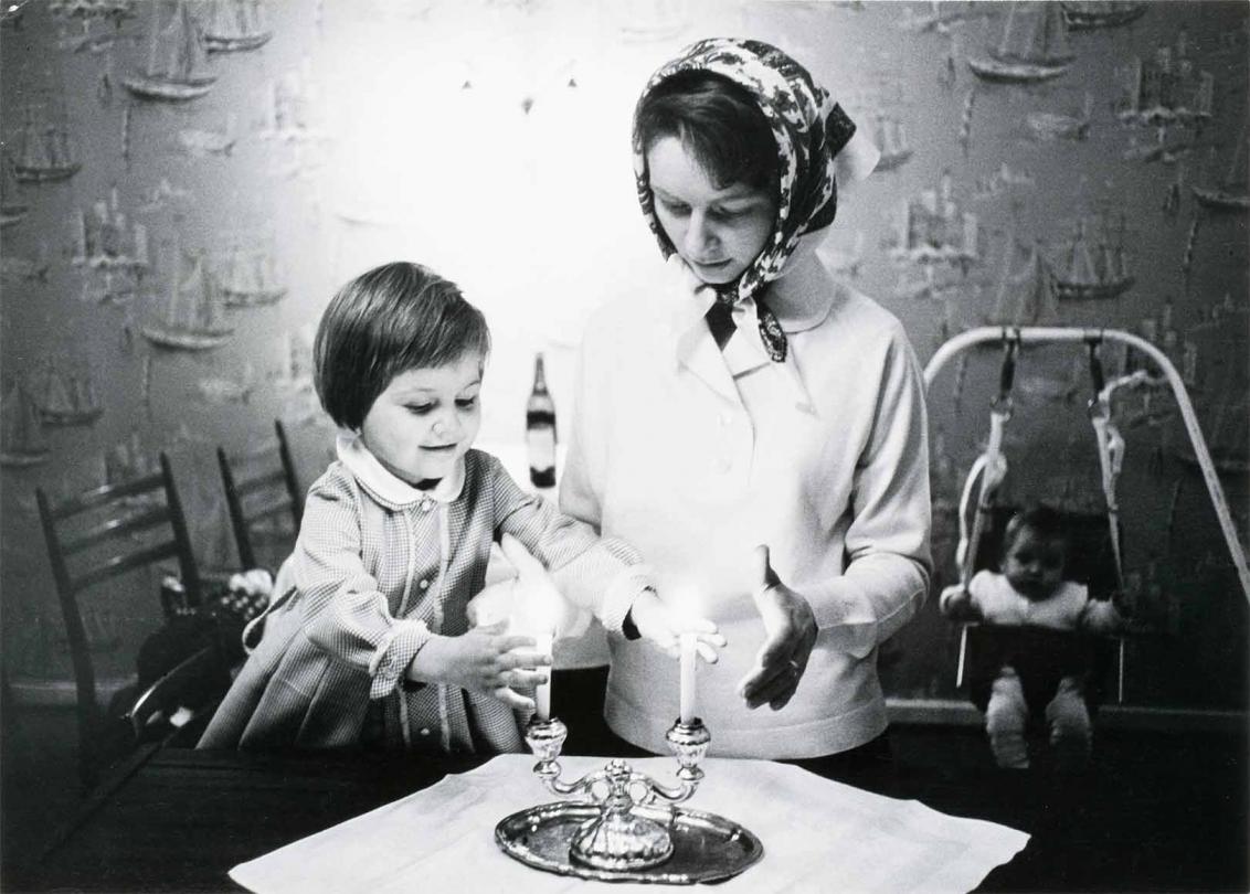 Black and white photograph: In the foreground, a girl and a young woman with a headscarf are blessing the Shabbat candles, which are placed in a two-armed candlestick on a table with a white tablecloth. In the background, a baby can be seen in a baby swing.