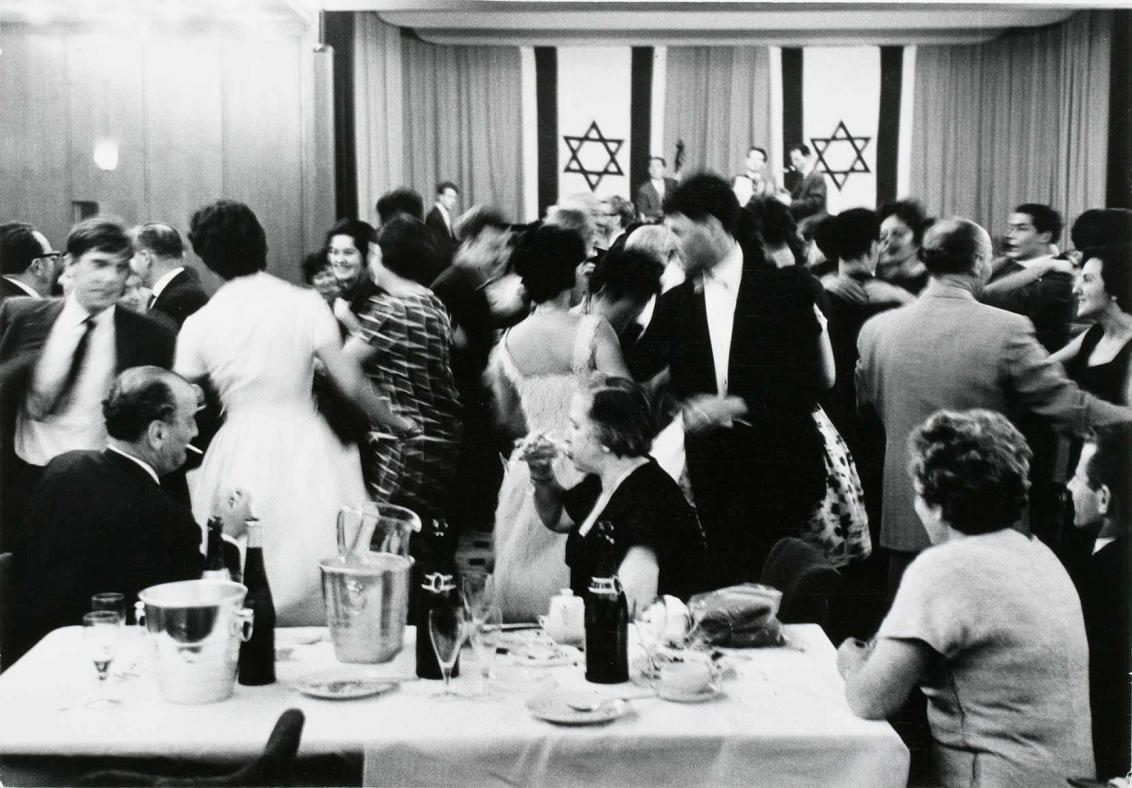 Black and white photograph: people dressed in festive outfits dancing; in the background is a stage decorated with two Israeli flags and a band playing.