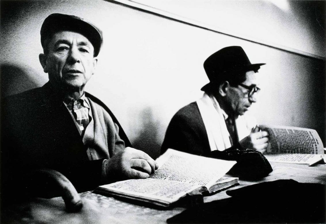 Black and white photograph: Two men with hats are sitting at a table in front of open books. The man on the left is looking towards the camera, while the man on the right is looking at the book and turning a page.