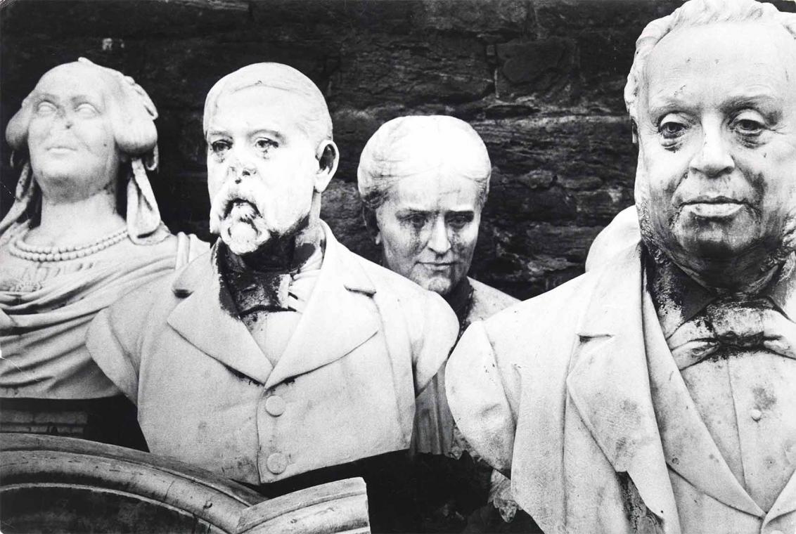 Black and white photograph: Two light-colored female and two male busts stand offset in front of a dark wall.