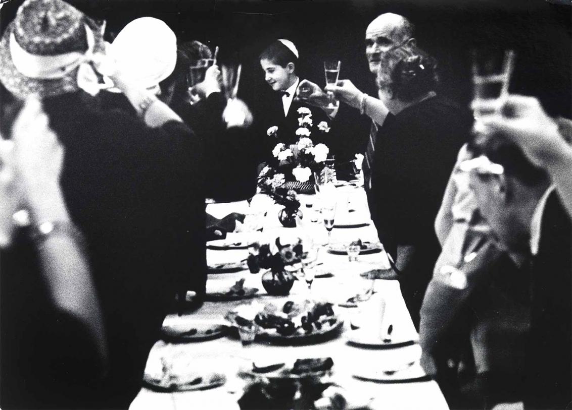 Black and white photograph: At a long, festively laid table, guests raise their glasses to greet a boy at the end of the table. He is wearing a light-colored kippah.