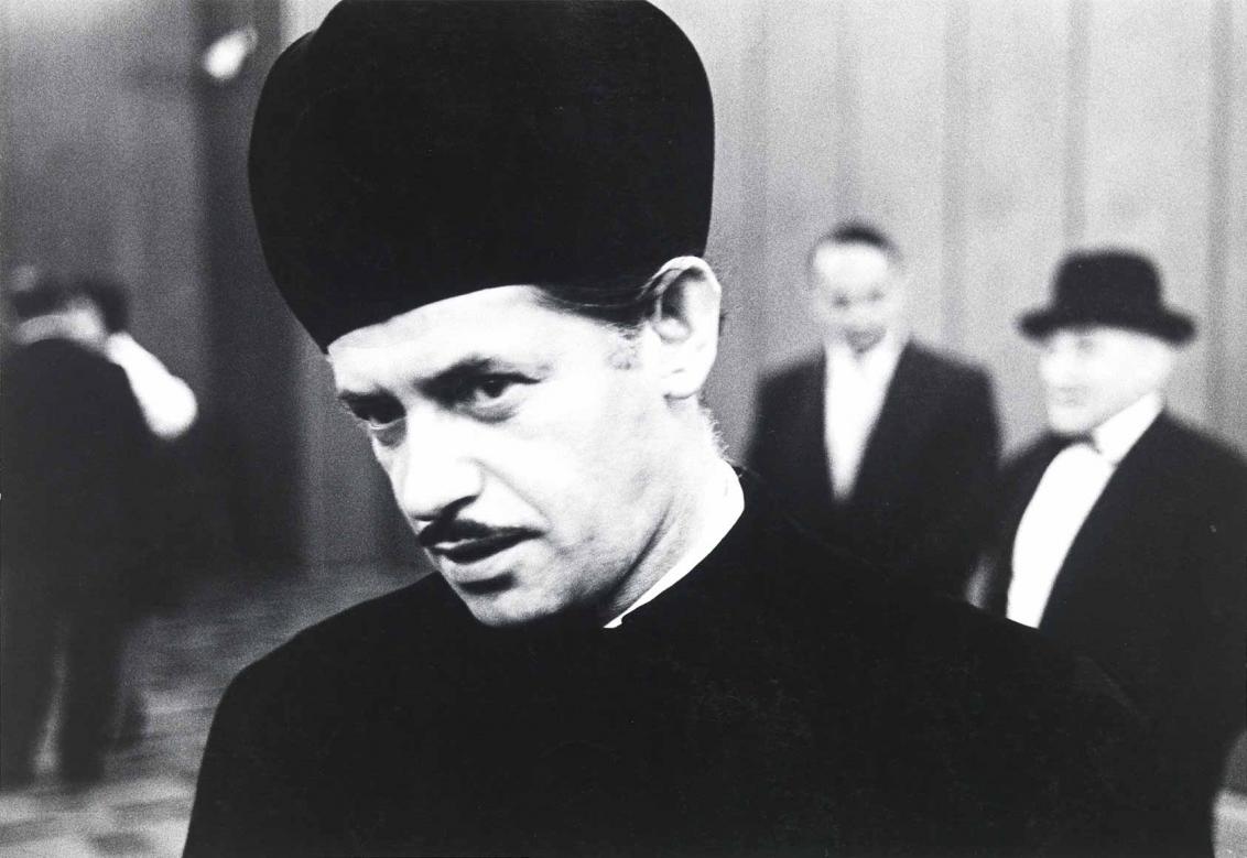 Black and white photograph: Portrait of a man with a narrowly shaved moustache. He is wearing the dark headgear of a cantor and dark clothing. Other people can be seen blurred in the background.