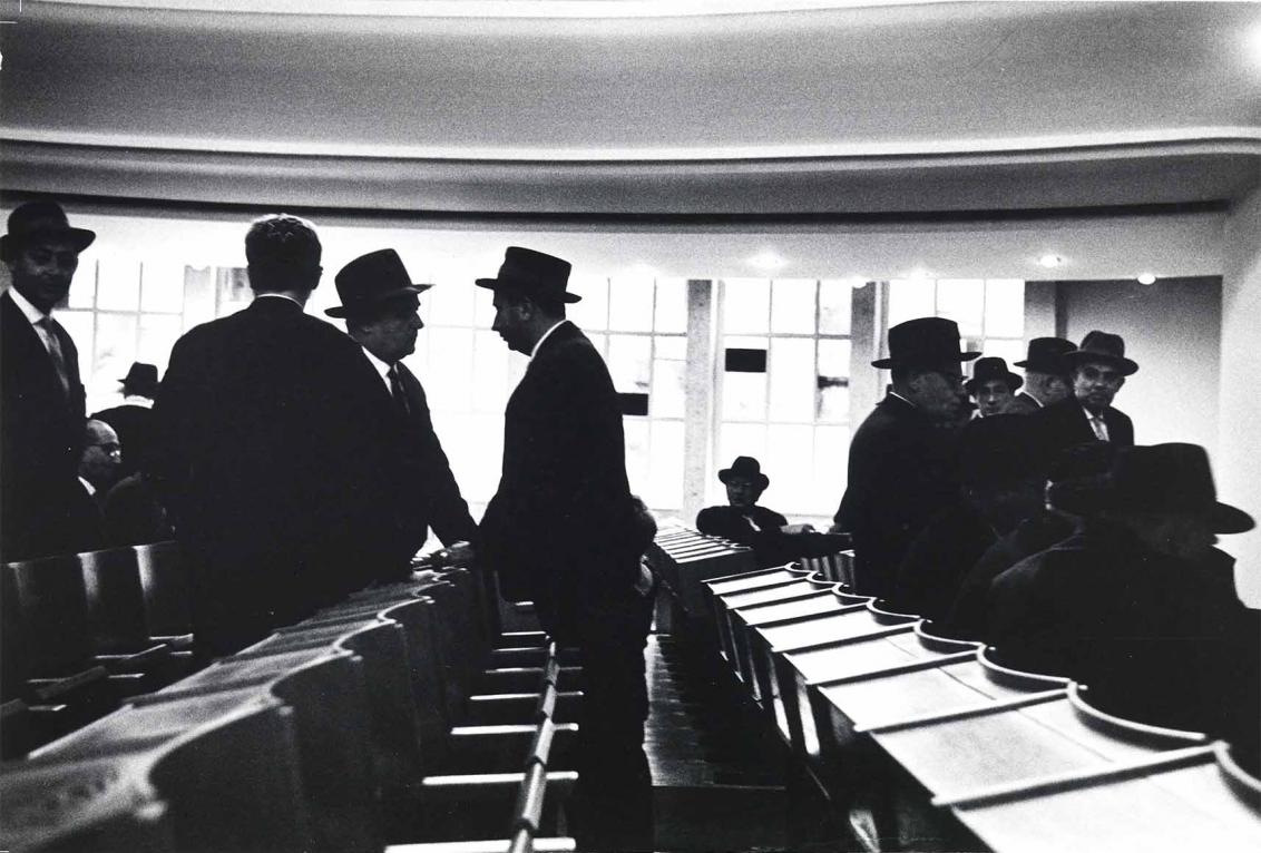 Black and white photograph: Some men stand and sit between and in the pews of a synagogue. They are wearing dark suits and various head coverings. In the background of the picture is a large, very bright window front. This makes the people in the foreground appear very dark and makes the photo very rich in contrast.