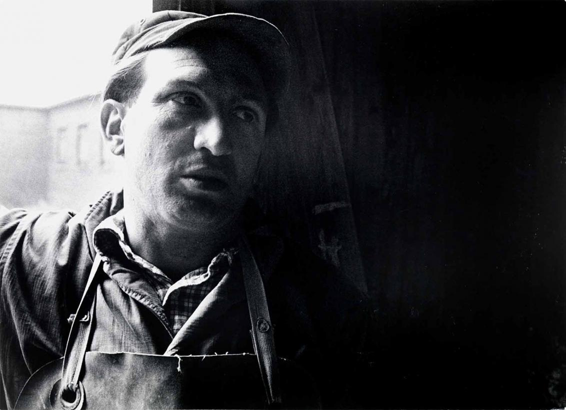 Black and white photograph: A man in a work shirt and apron stands in front of a window or door frame. Only his head and shoulders are shown. Light is shining on his face from the left and he is looking to the right.