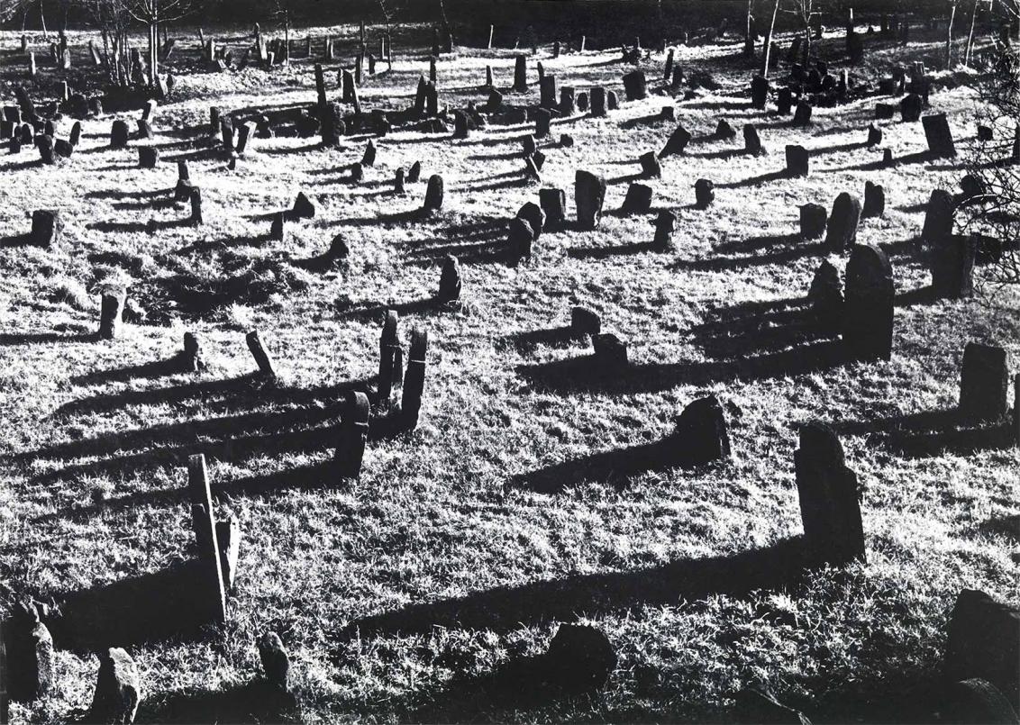 Black and white photograph: A large number of gravestones stand on an area overgrown with grass. Most of the stones are not upright, but leaning forwards or backwards. The sun is shining so that the gravestones cast long shadows.