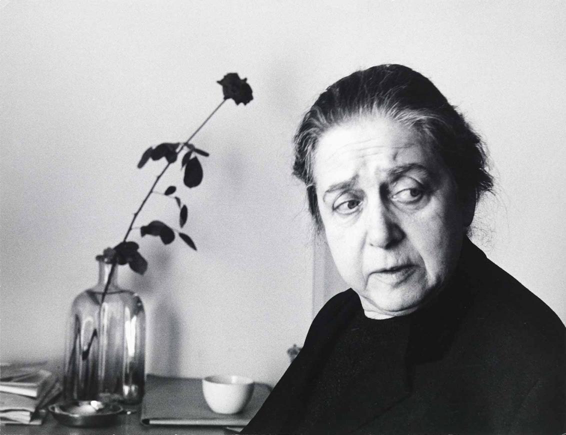 Black and white photograph: Portrait of an older woman with dark, graying hair tied back. She is looking to the right. To her left is a rose in a glass bottle on a table on which a cup and a few notebooks can also be seen.