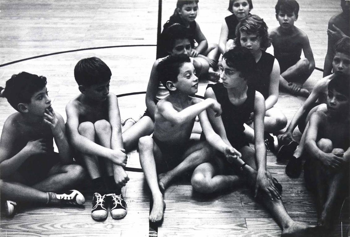 Schwarzweiß-Fotografie: Mehrere Kinder sitzen in einer Gruppe zusammen auf dem Boden einer Turnhalle. Sie tragen alle Sporthosen und teilweise Sportschuhe, die Jungs tragen keine Oberteile, die Mädchen tragen ärmellose Oberteile.