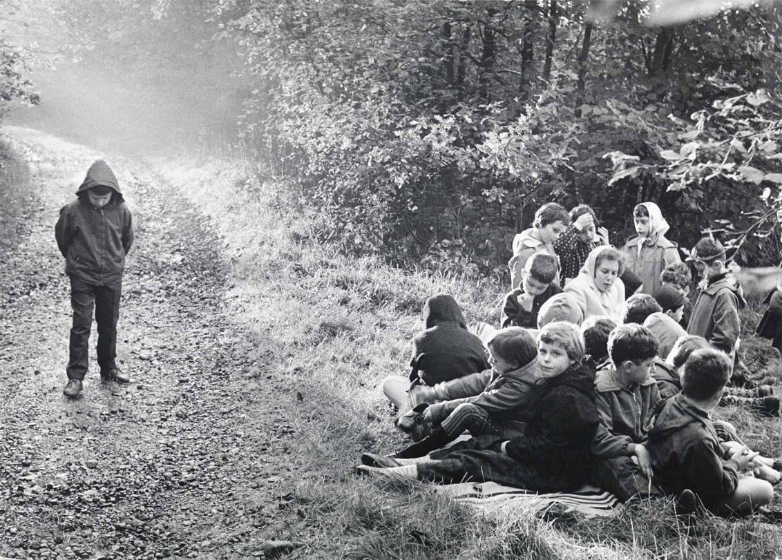 Black and white photograph: A group of children sit and stand close together on the right next to a forest path. An adult woman sits in the middle. On the forest path, slightly away from the group, stands a boy. He has his hood up, his head down and his hands clasped behind his back.