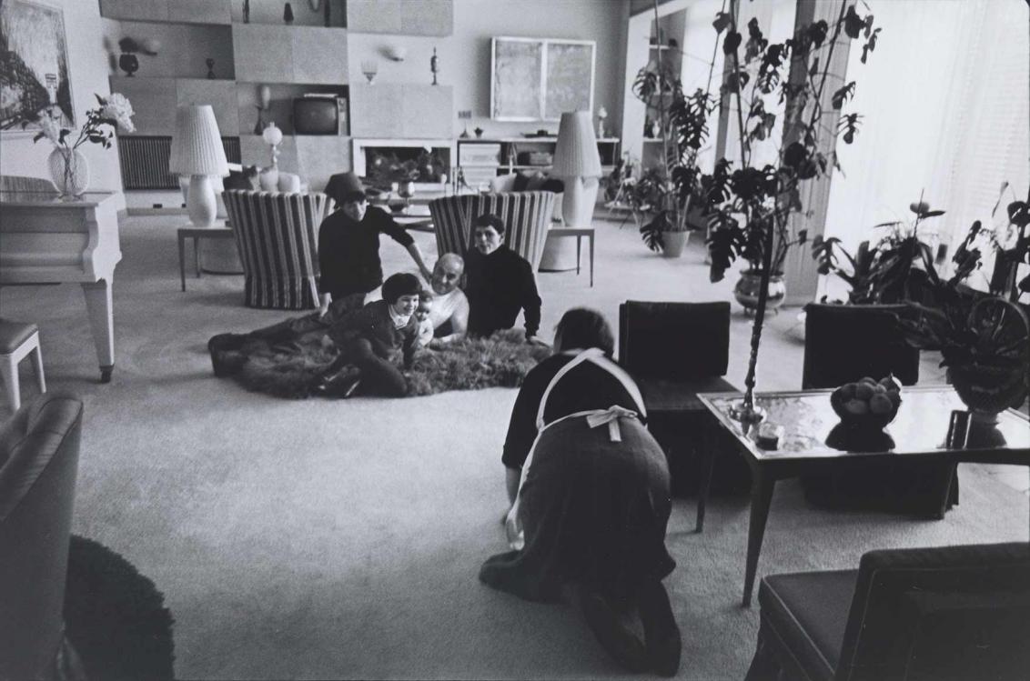 Black and white photograph: A family with children is sitting on a dark fur rug in a large living room. An elderly lady wearing an apron squats on all fours in front of the people on the fur and looks at them.