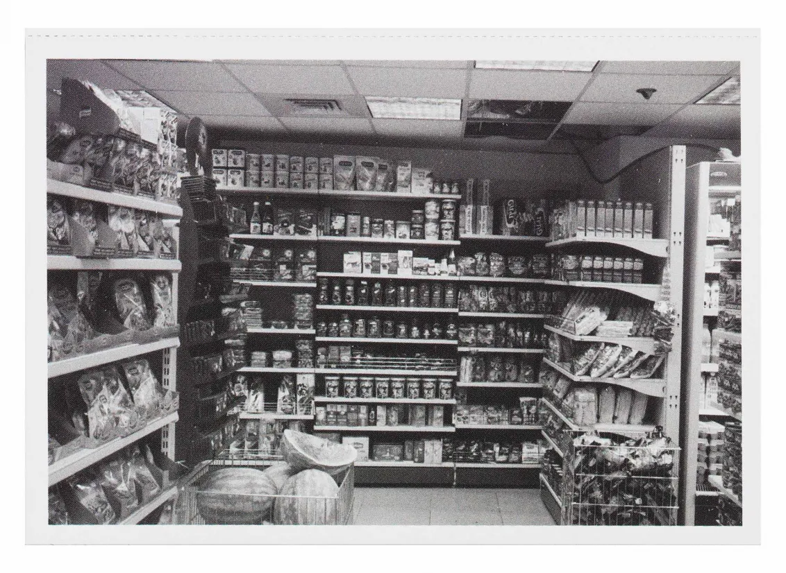 Black and white photograph of a grocery store aisle, a pile of watermelons is surrounded by boxes of food and snacks that line the shelves