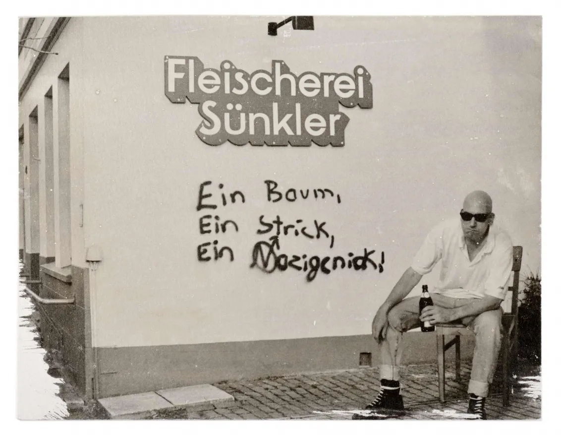 A man wearing sunglasses and holding a beer sits next to a wall with graffiti