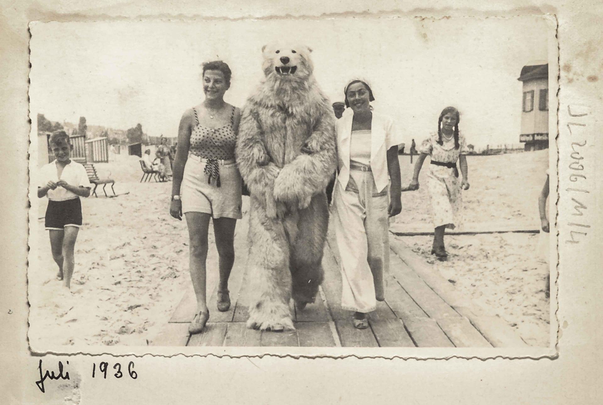 B-W photo: Two summery dressed women have hooked a person with a polar bear person in their midst and are walking towards the camera on a wooden walkway on the beach.