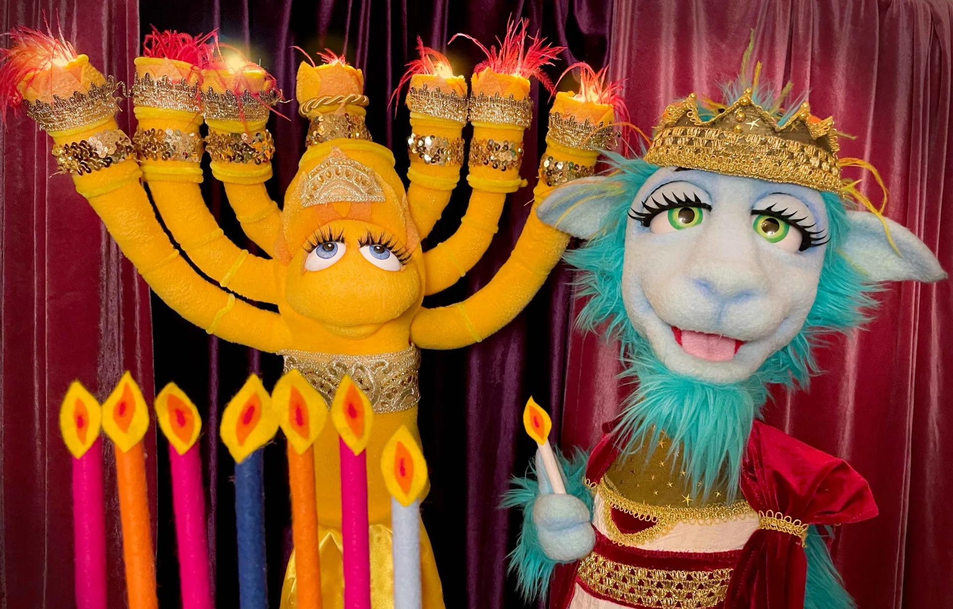 Two hand puppets, one in the shape of a Hanukkia and one as a sheep, stand in front of a red stage curtain with eight candles.
