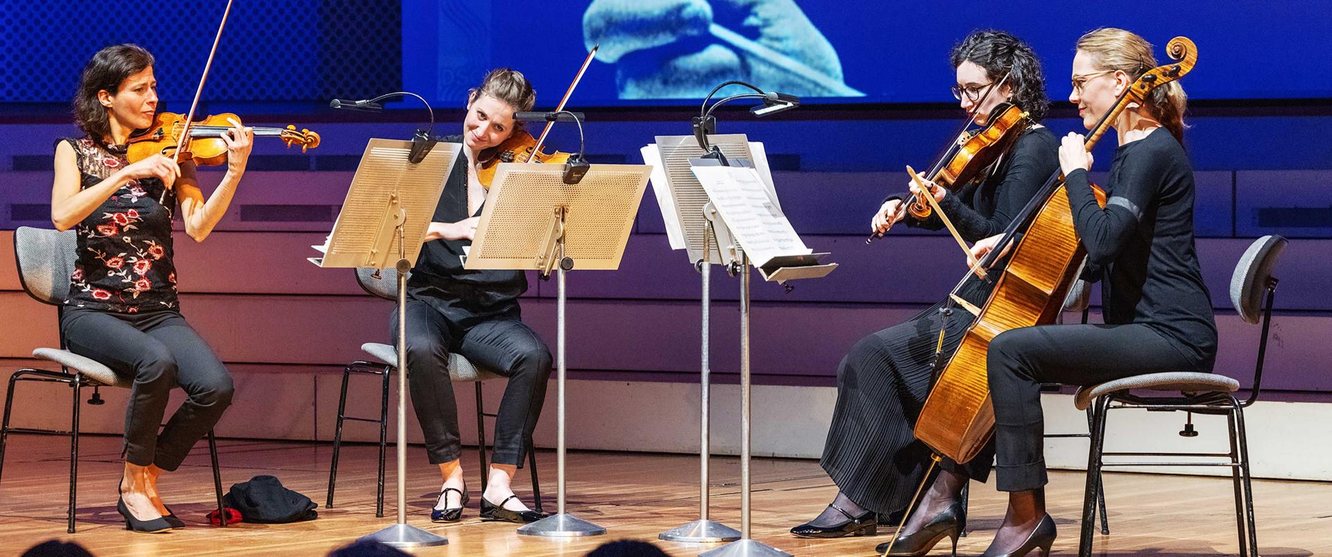 Four women are playing their instrumnets in a quartett constellatin. Three of them play the violine, one plays a cello.