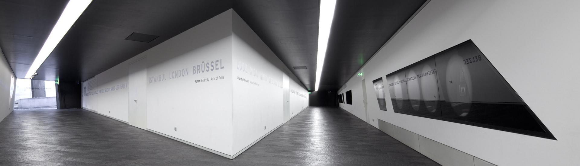 Two corridors with dark slate floors and white walls; one leading to the right featuring a display cabinet; the other leading to the left towards a door to the Garden of Exile; the walls between the corridors feature city names like Istanbul or London