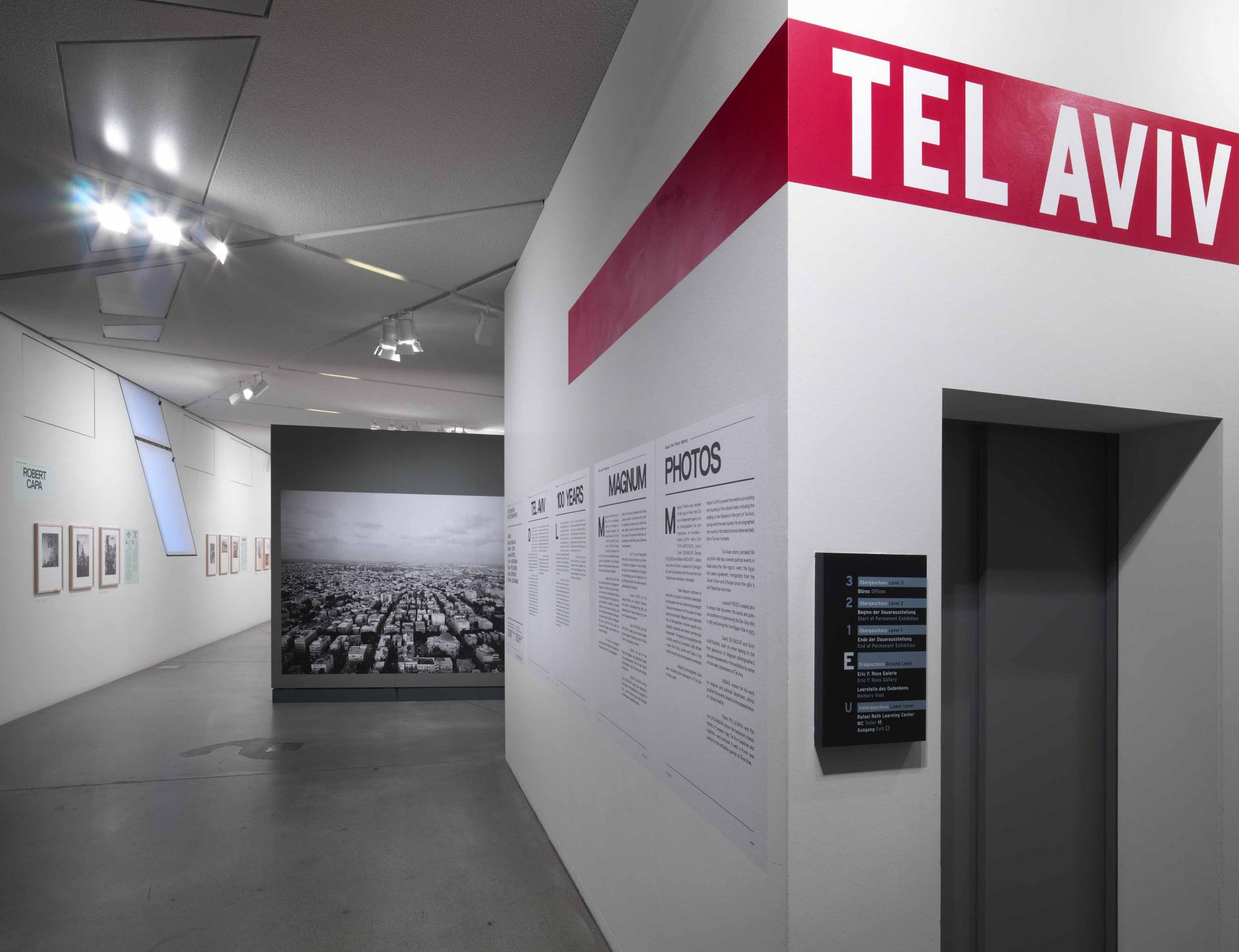 Blick in den Eingangsbereich der Ausstellung: Rechts eine Wand mit Beschriftung, im Hintergrund ein großen Schwarz-Weiß-Foto, das ein Lufbild einer Großstadt zeigt