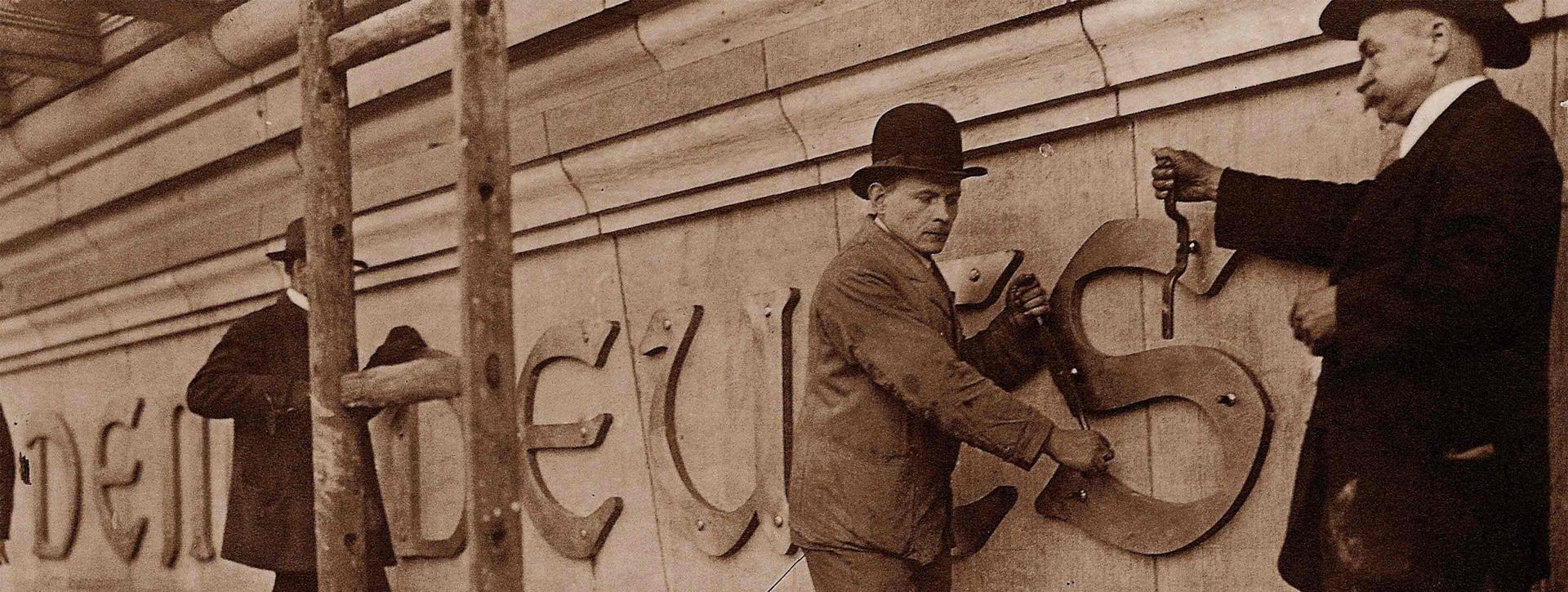 Craftsmen with tools apply lettering to a facade.