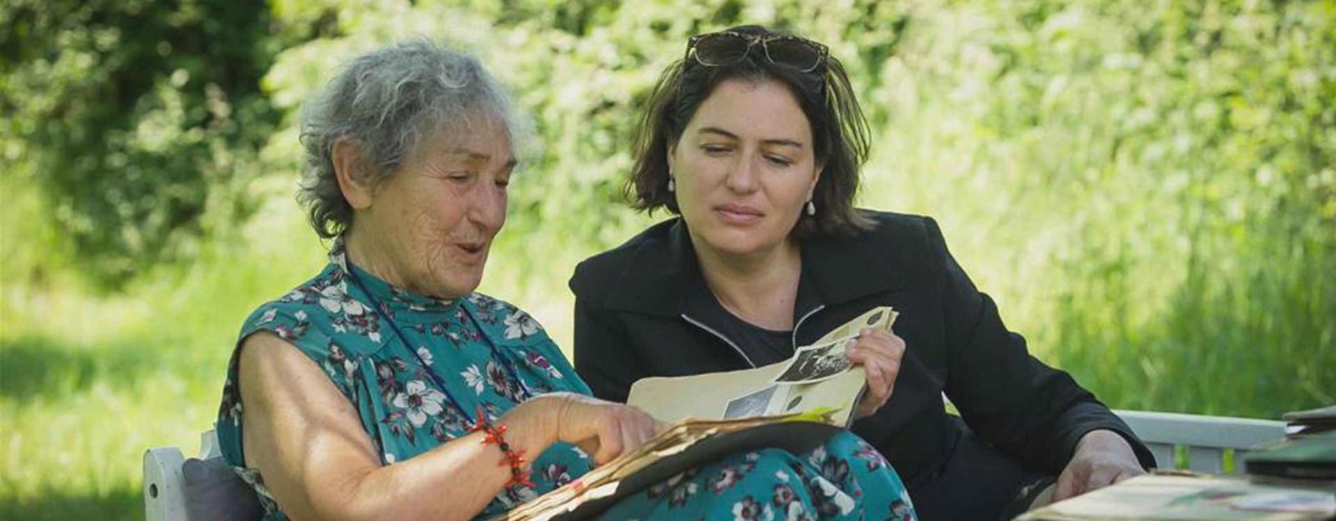 Two women sit on a garden bench and leaf through a photo album together.