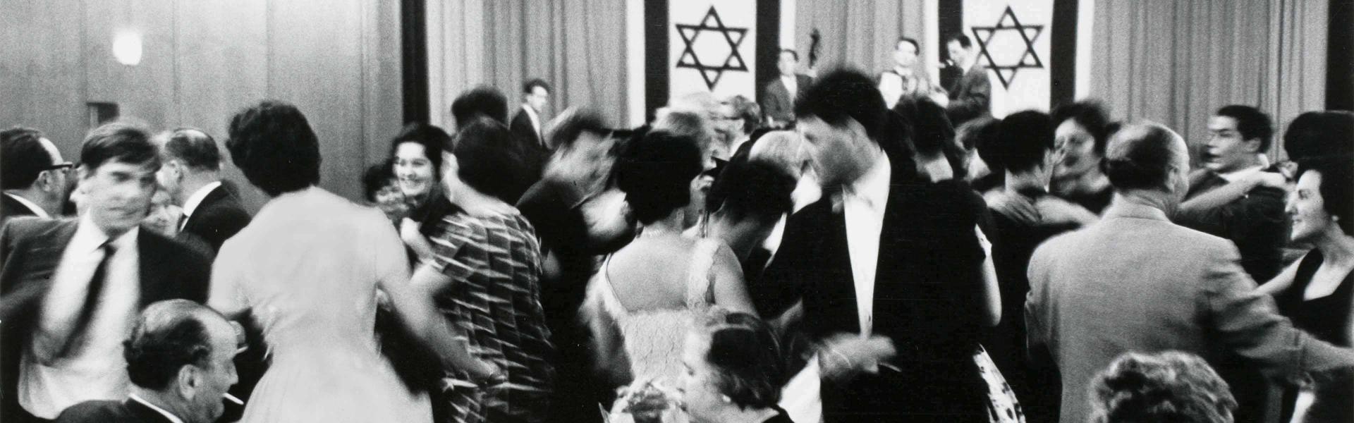 Black and white photograph: people dressed in festive outfits dancing; in the background is a stage decorated with two Israeli flags and a band playing.