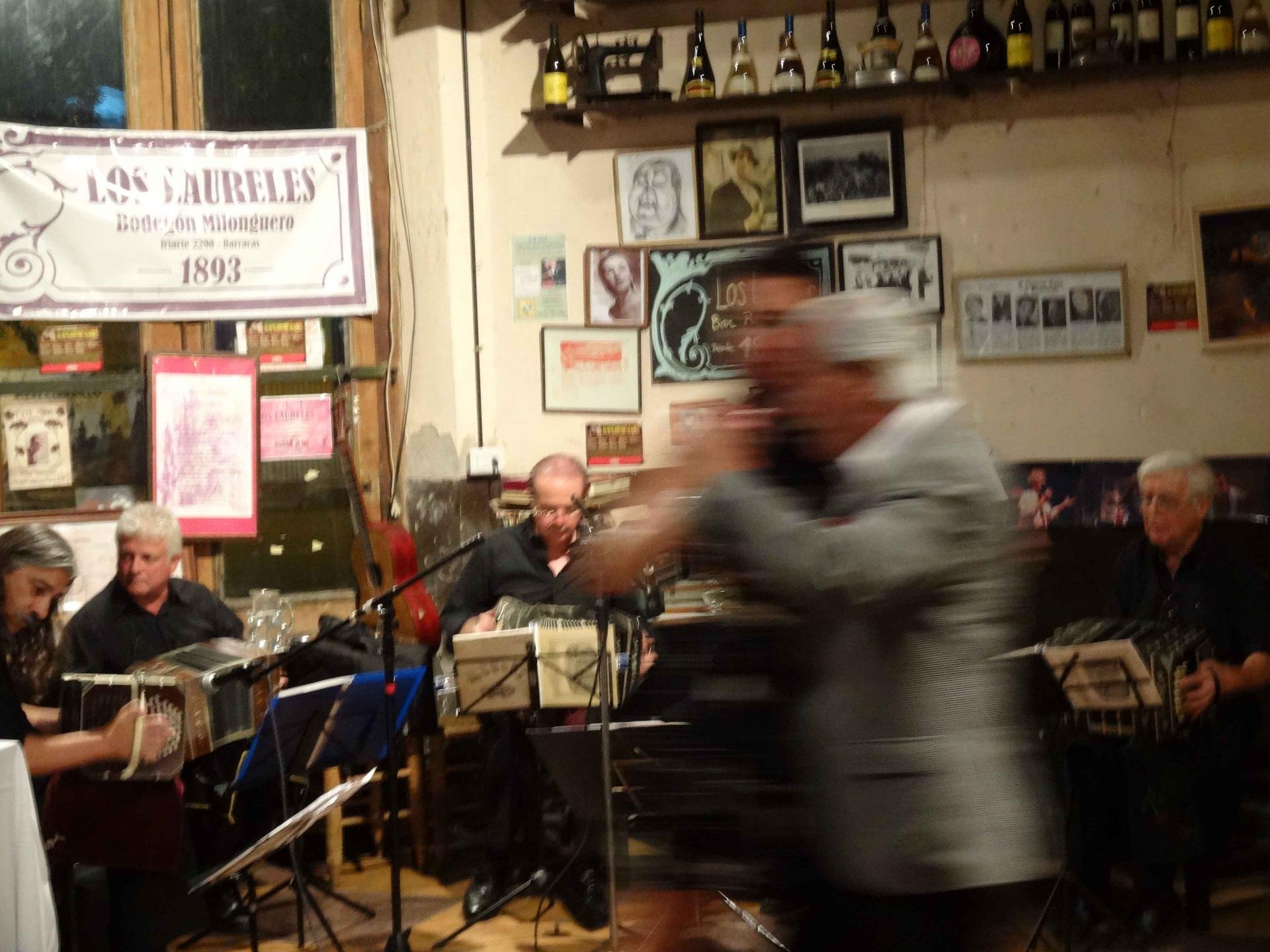 View into a dance hall. A couple dancing in the foreground out of focus, musicians sitting behind microphones and music stands in the background.