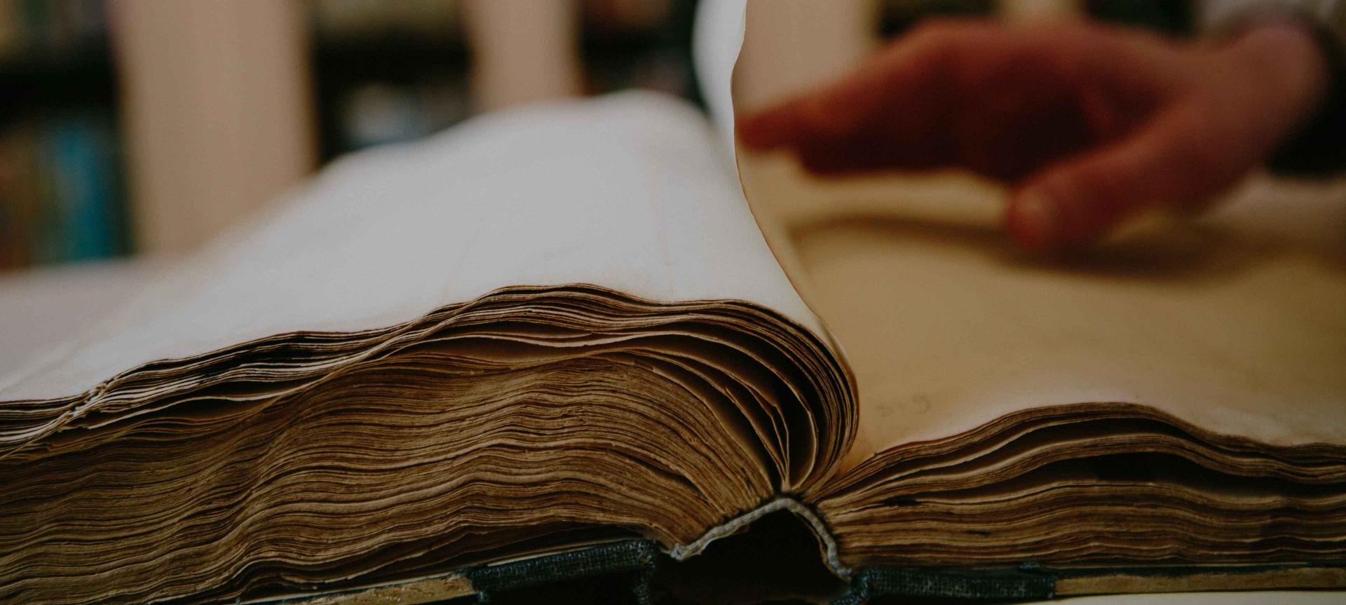 Hand turns over a page of an open thick, old book.