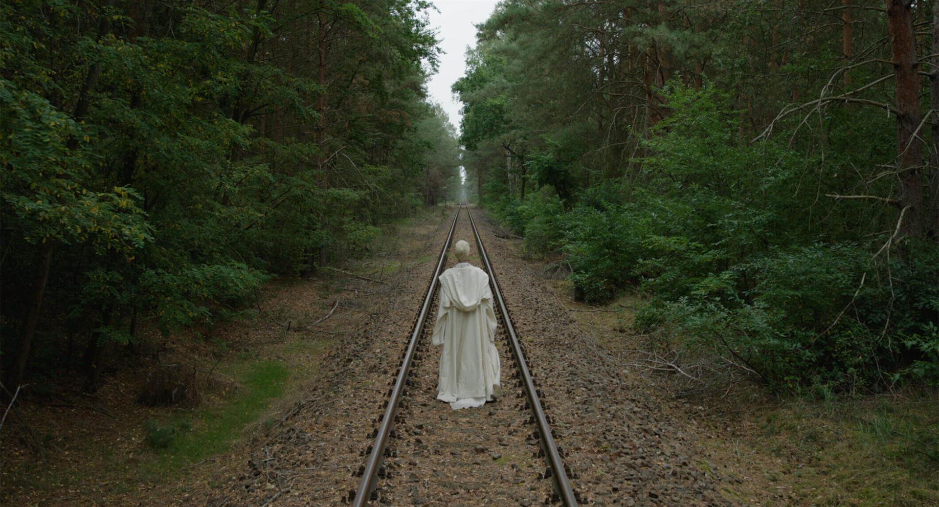 The film still shows a person from behind, dressed in a white frock. The person follows railroad tracks that run dead straight through a forest