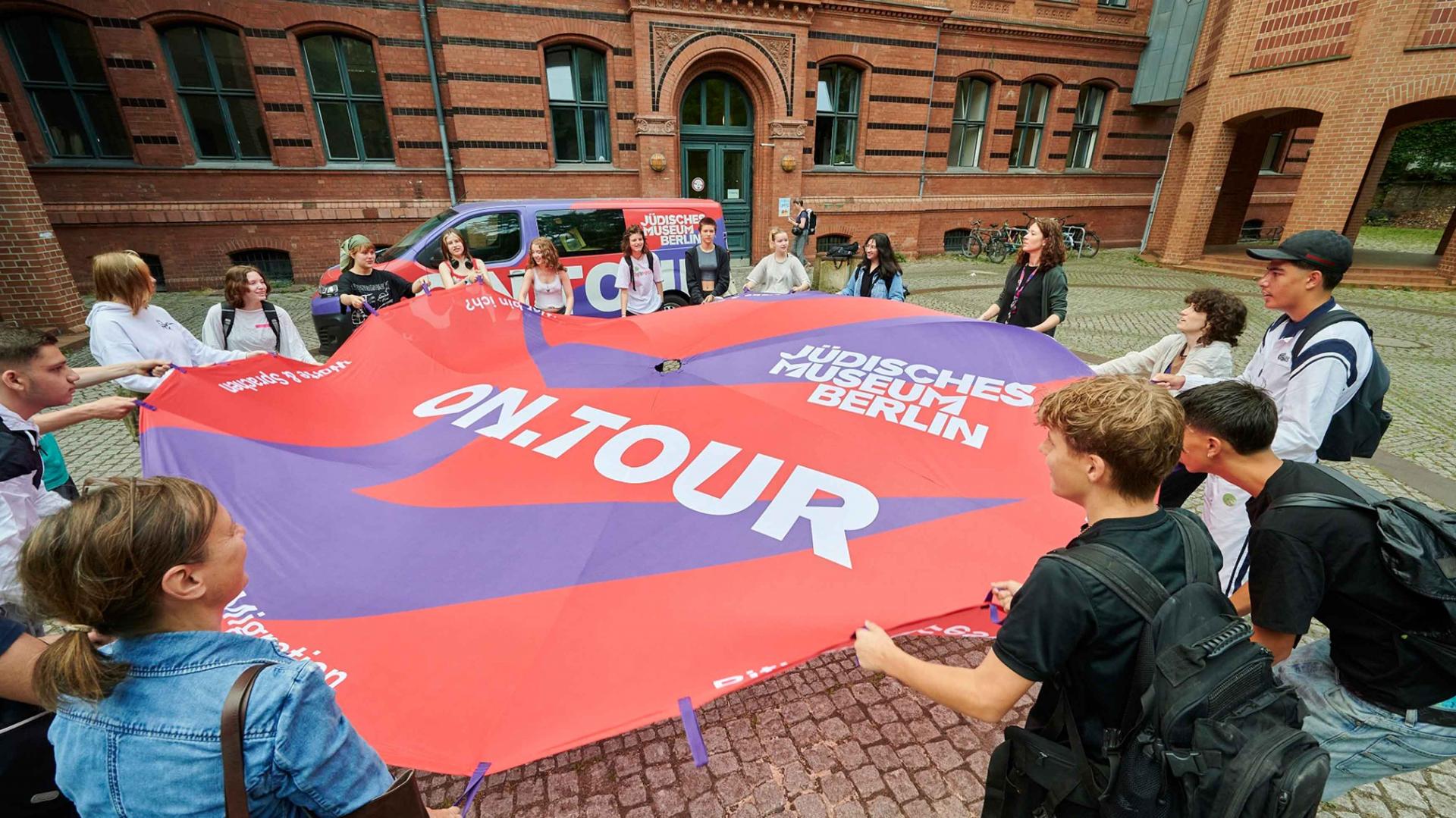 A group of young people stretch out a large cloth in a courtyard with the words on.tour and Jewish Museum Berlin on it.