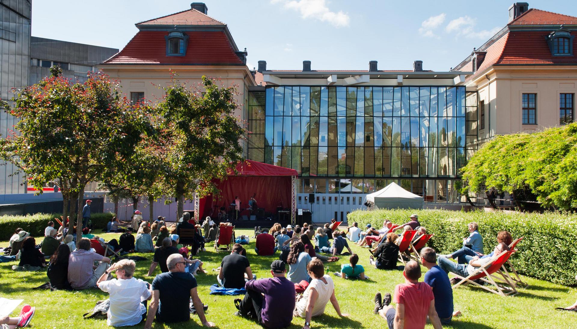 Foto vom Sommerfest im Garten des Museums. Besucher*innen sitzen in der Sonne auf dem Rasen und schauen in Richtung einer kleinen Bühne.