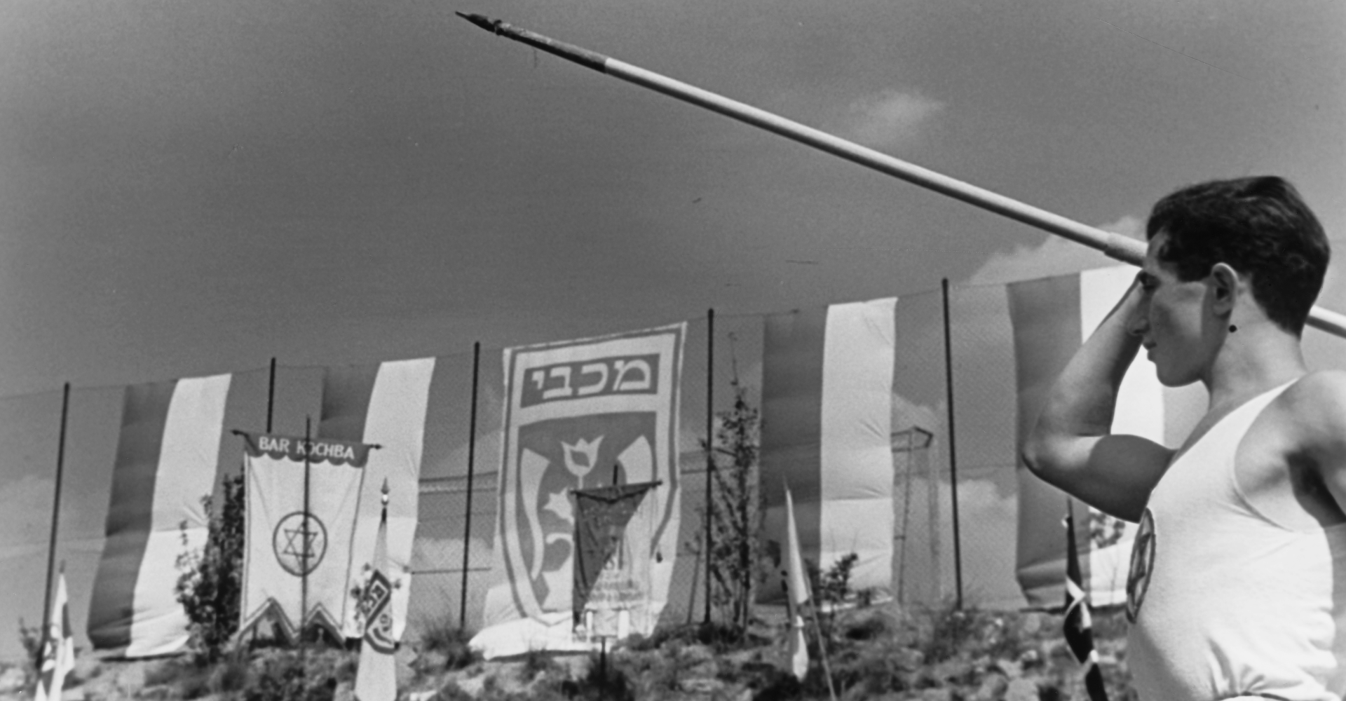 Young man holding a spear in a throwing position above his head. In the background flags.