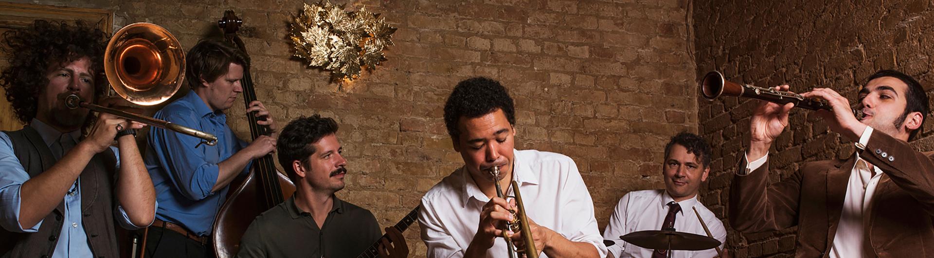 Six male members of a jazz combo play their insturments in front of a brick wall.